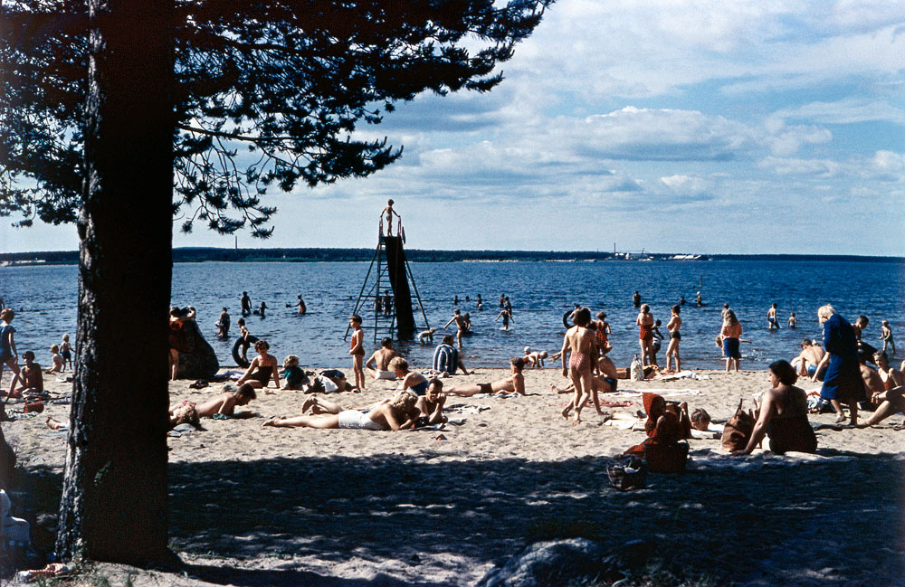 Vy över badstrand med mycket folk i badkläder.