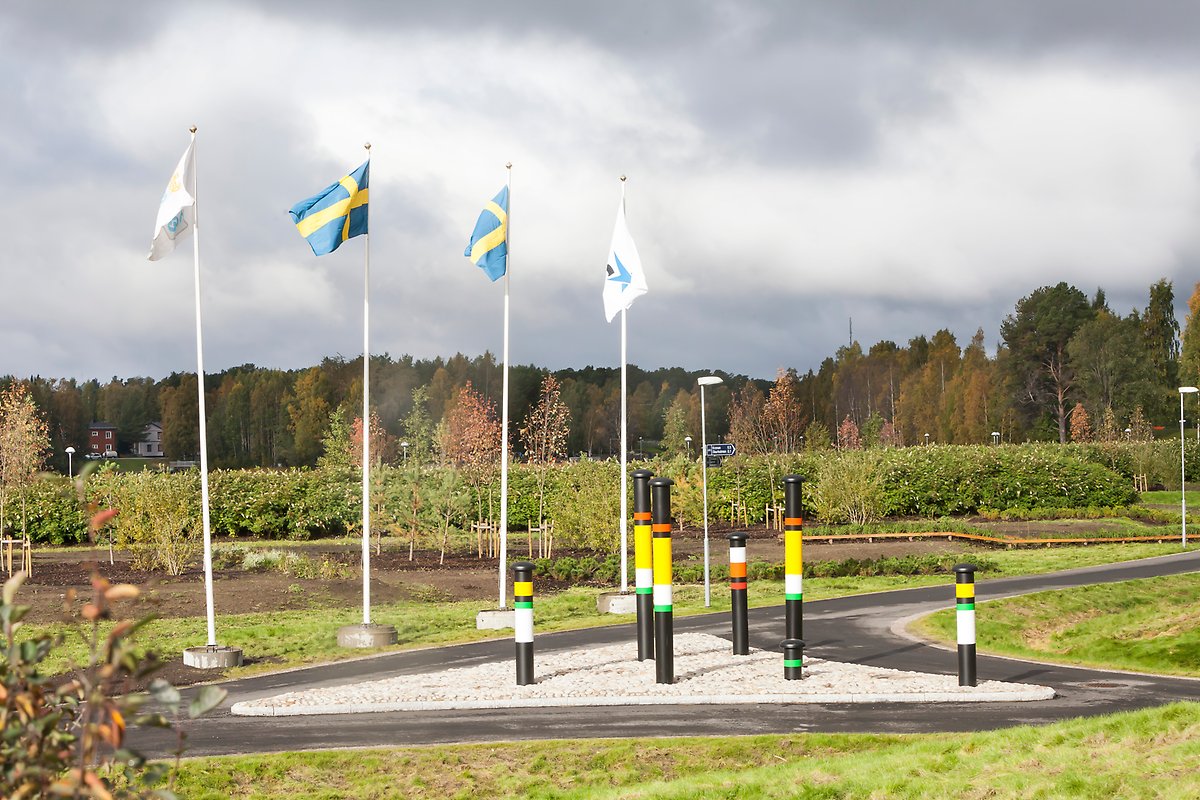 Flaggstänger med svenska flaggan hissade. Framför dem syns en trekantig sandyta med ventilationsrör från vatten och avloppsanläggningar. De svarta rören är målad i vitt, grönt, gult och rött.