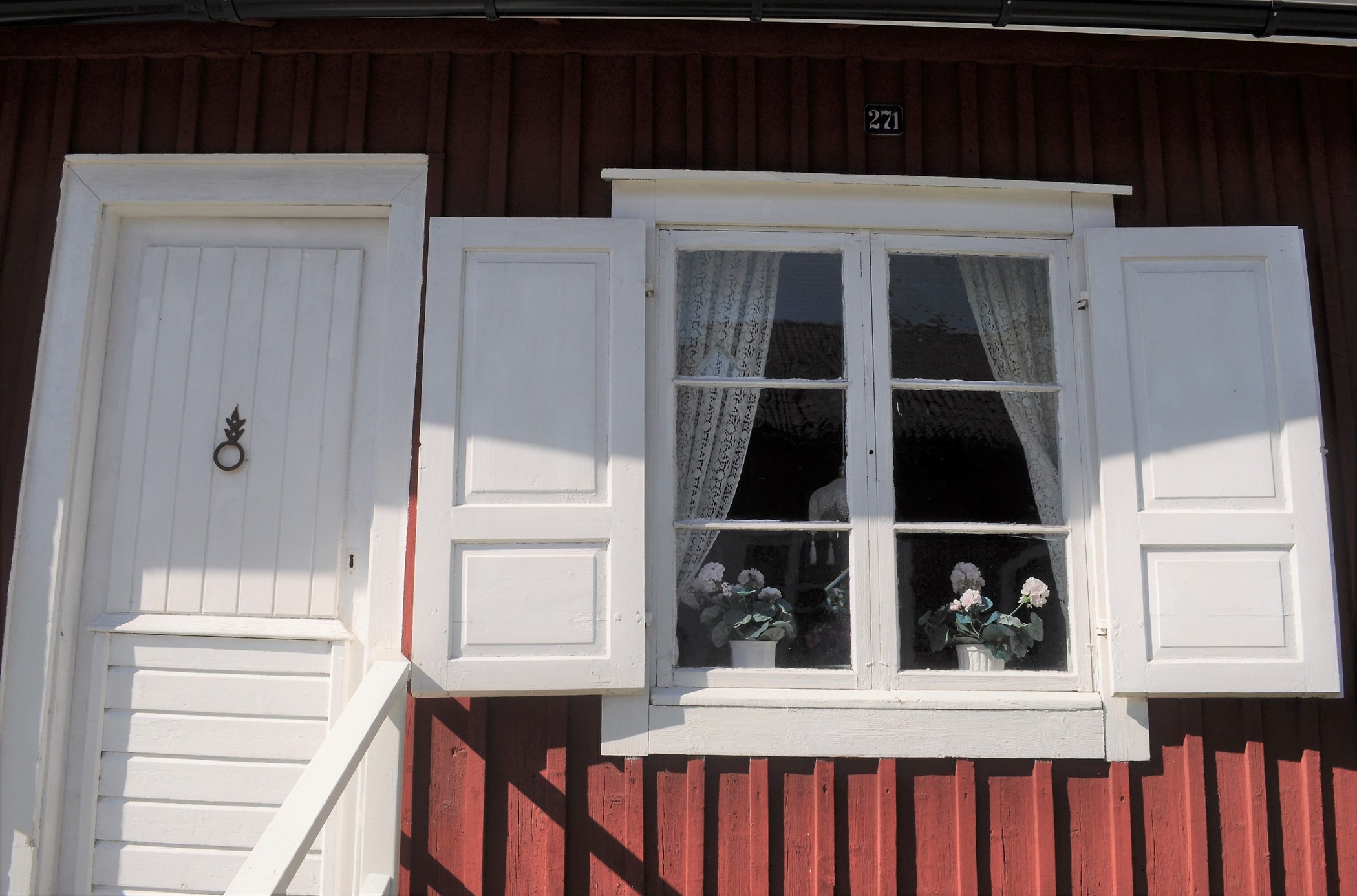 Vitt fönster och vita fönsterluckor och röd träfasad. Vita blommor och spetsgardiner i fönstret.