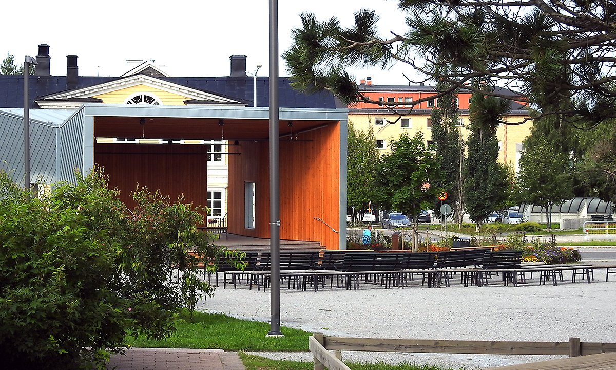Stadsparken väg mot scenen med träd i förgrunden. Belysningsstolpar är riktade mot träden
